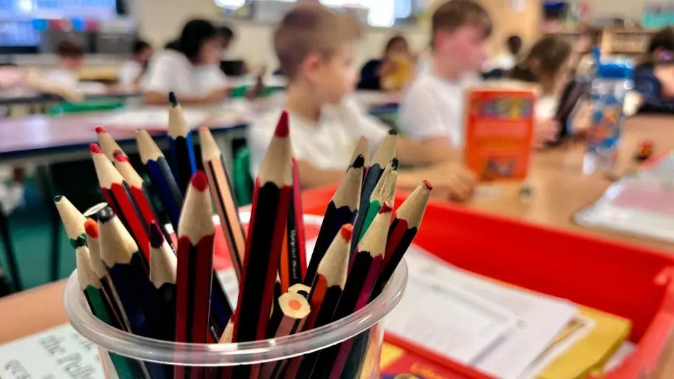 A pot of pencils is in focus in the foreground of the picture. They are on a school teachers desk with a bright red in-tray. In the background there are children sitting in rows at desks but they are…