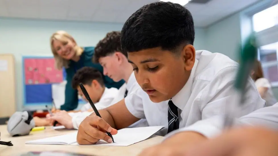 A medium shot of teenage boys in school uniforms sit side by side at their desks, working on a classroom task. The students write in their notebooks while smiling. The classroom environment is bright…