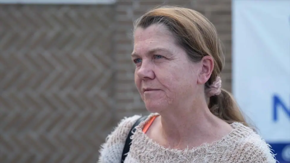 A woman with brown hair looking off screen, she has her hair in a ponytail and is wearing a light brown jumper.