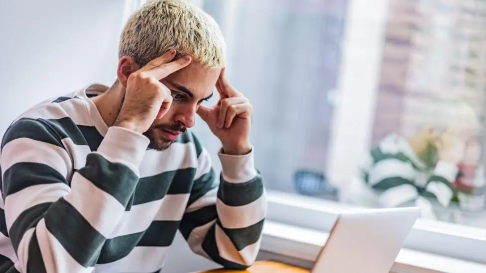 A young man with short bleached blonde hair and a beard, wearing a striped long-sleeved T-shirt, sits at a desk, looking toward an open laptop. He looks frustrated and is resting his fingers against…