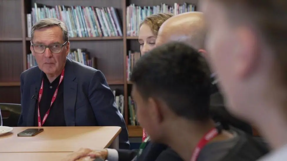 Alan Milburn author of the report sits on the left at the end of the table facing students in conversation, he wears a navy suit, red lanyard and black polo shirt. He looks serious in deep…