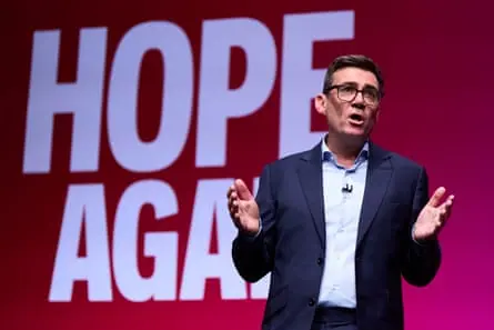 Andy Burnham speaks at the conference. He has his hands raised and outstretched, and stands in front of white lettering reading Hope Again on a pinkish red background. He wears glasses and a dark…