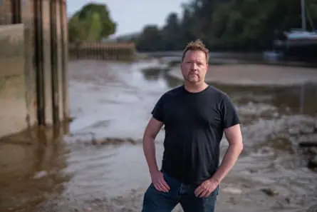 Rob Worthington stands in mud at the edge of the river. He has light brown hair and a short beard, and wears a black T-shirt and jeans.