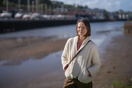 Emma Roberts stands by the side of the river with boats moored on the other side in the background. It is low tide and there are mudbanks.