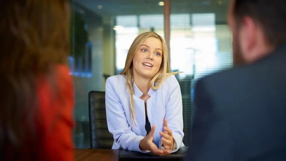 Woman sits at a table in an office being interviewed by a man and a woman whose blurred backs are in the foreground.