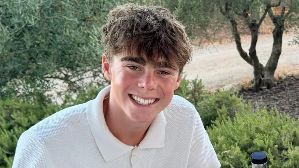 Hugo Thompson smiles at the camera. He is wearing a white polo shirt and sat in front of olive trees in a field