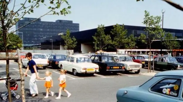 This is a grainy 1970s photo showing a large angular black shopping centre and some small newly planted trees. In the foreground is a largely full car park and a nadult pushing a trolley while two…