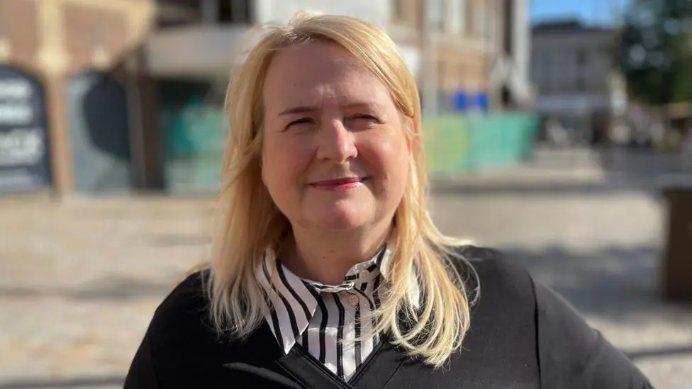 This is a head and shoulders picture of Dr Kardi Somerfield looking at the camera and smiling. The background is blurred but she is standing in a town centre.