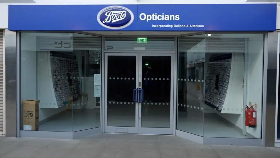 This a picture of a shop front for a branch of Boots Opticians. The blue sign is above two large windows with glass double doors in the middle. The shop is closed.