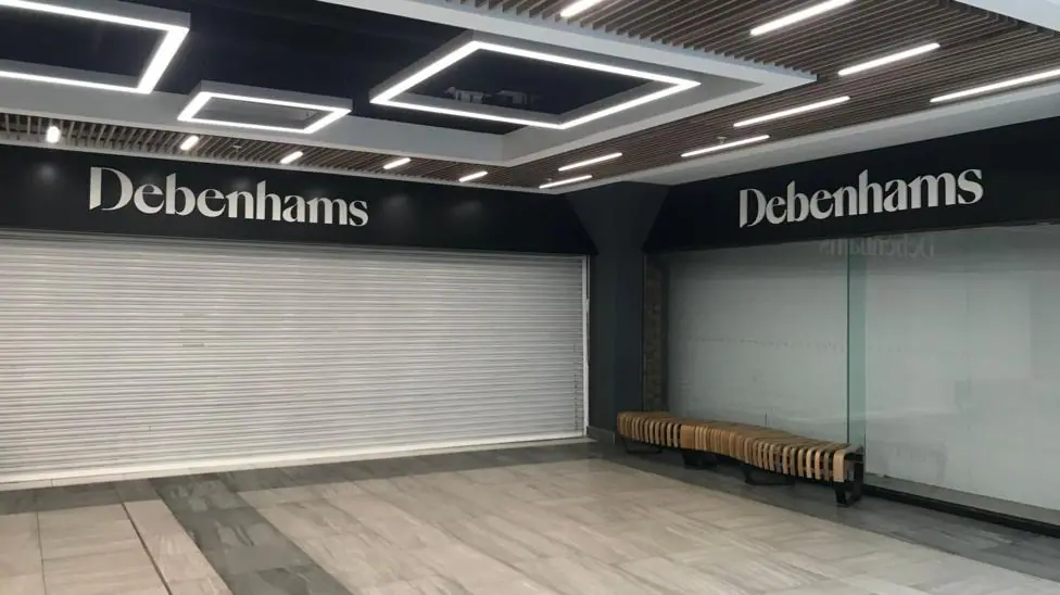 A shot inside a shopping centre shows shuttered windows and Debenhams store signage.