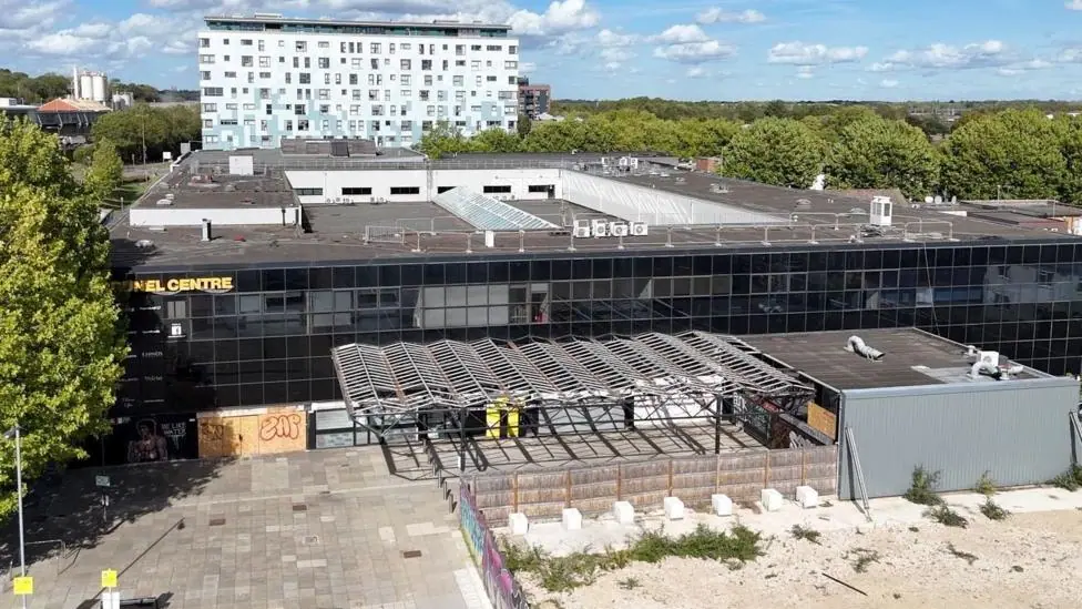 This is an aerial picture of a large angular black shopping centre. There are trees to the left and right and in the distance behind the centre is a large high rise block. There is a blue sky with…