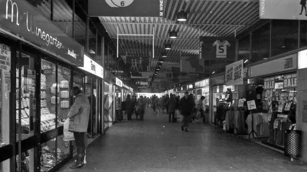 This is a black and white photo of the inside of a shopping arcade. Shops line each side of a walkway with people seen browsing in the windows.