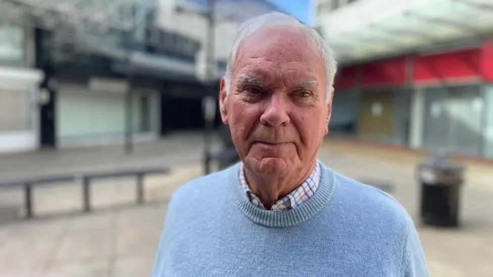Keith Ely stands in a deserted shopping centre, which is out of focus behind him. He is wearing a light blue crew-neck sweater over a white, brown and blue checked shirt. He has white hair and is…