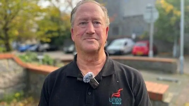 Vic Fraser has short, grey hair and is wearing a black T-shirt with Unite the Union written on it with the Unite logo. He has a small microphone attached to his T-shirt and there are cars parked…