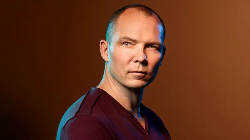 A headshot of Jonathan Blow in a dark v neck top against a dark background, with a shaved head, staring off camera