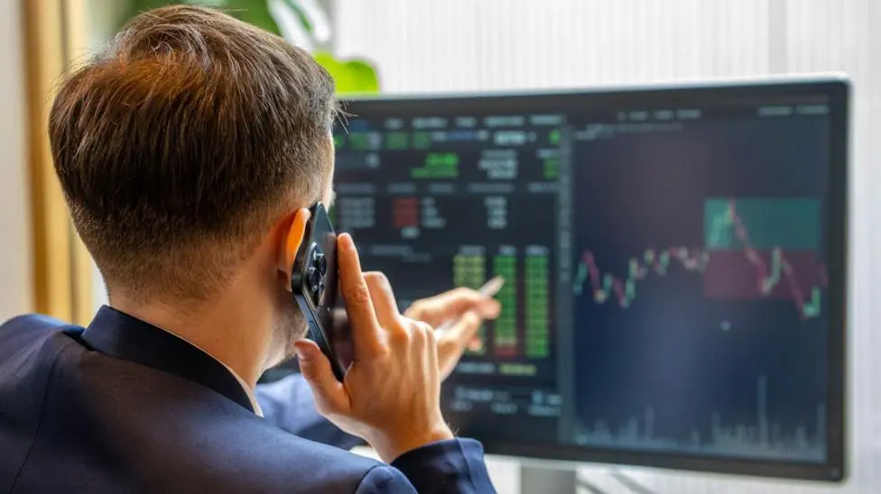 A man holds a mobile phone to his ear while pointing at a large computer screen that is displaying charts and numbers.