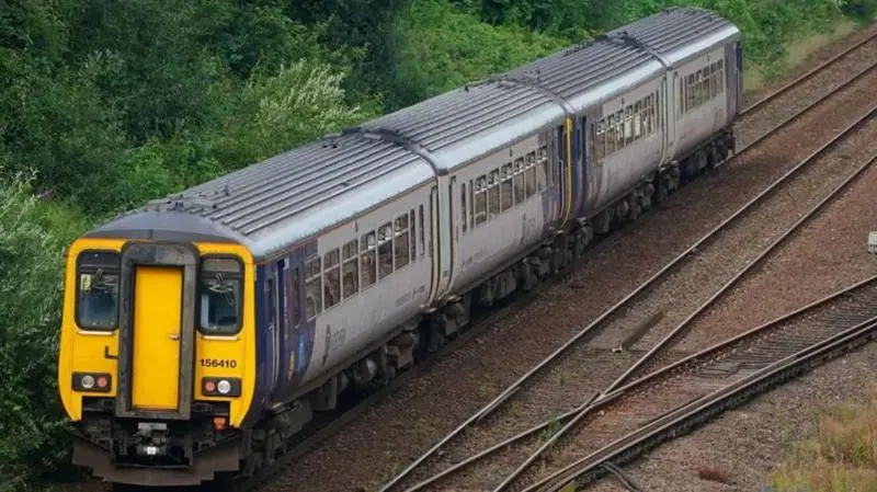 A two-car Northern passenger train travels along a railway line bordered by greenery, viewed from an elevated position, with multiple tracks and points visible beside the train.