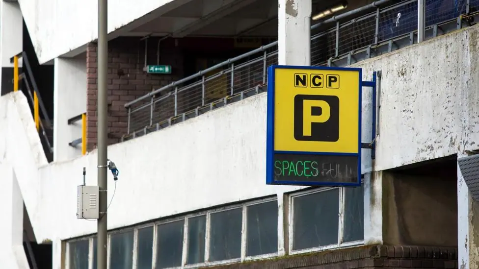 The exterior of a multi-story car park is seen with a sign the has the 'P' logo indicating parking, with the letters NCP above and 'spaces' below, taken in London in 2021.