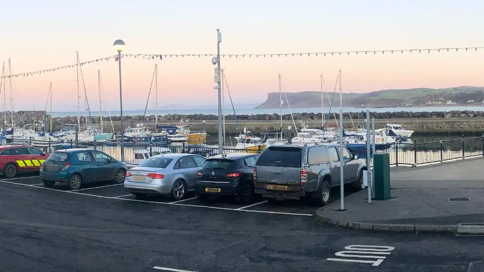 Car park in Ballycastle with a number of cars parked alongside marina, boats, water and mountains are visible in the background, the sky is at sunset and there are string lights attached on poles…
