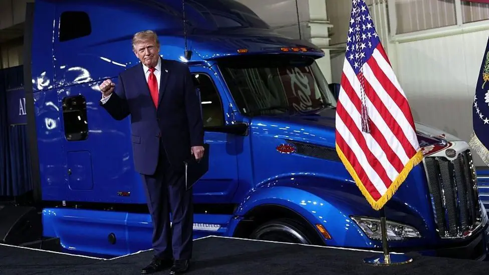 Donald Trump stands in front of a shiny blue truck and next to an American flag holding a raised fist during a visit to the Peterbilt Motors truck plant in Texas.