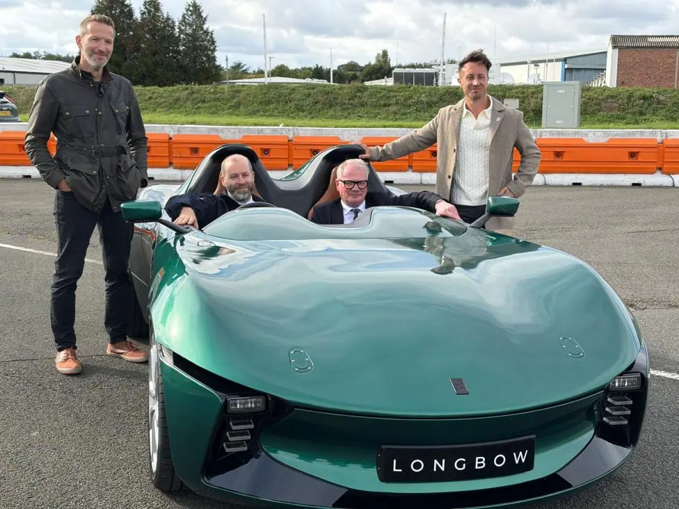 Two men stand beside a blue sports car with a “Longbow” number plate, while two others sit inside on a test track. An orange-and-white barrier and greenery are visible in the background.