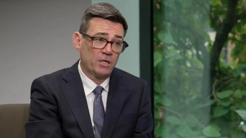 Andy Burnham, who has short dark hair and black glasses, wears a white shirt and a navy blazer
