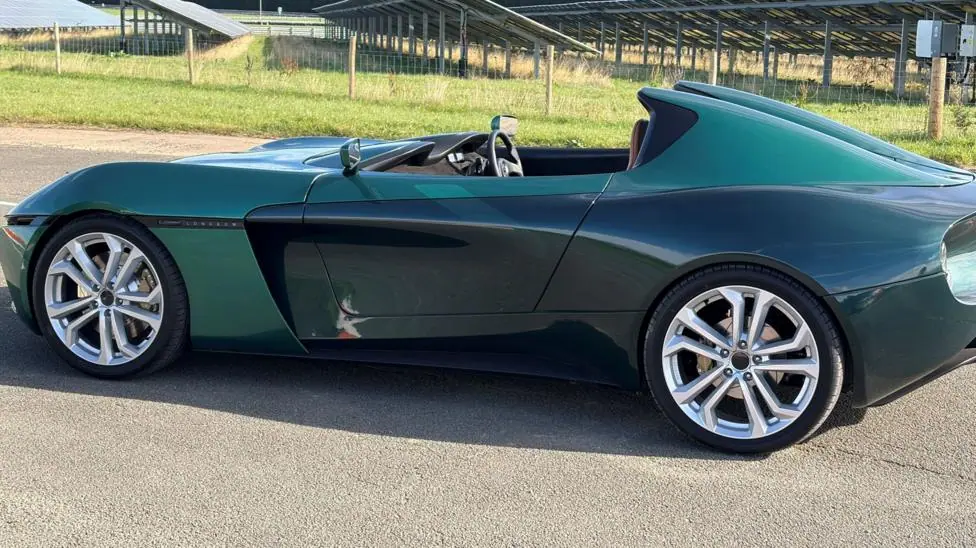 A long blue sports car pictured from the side. Some grass and some metal slates are in the background.