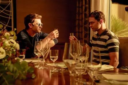 Two men sit at a dining table facing each other and raising a glass