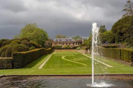 View of large country house across a manicured lawn with neat hedges to each side and a pool with spurting fountain in the foreground.