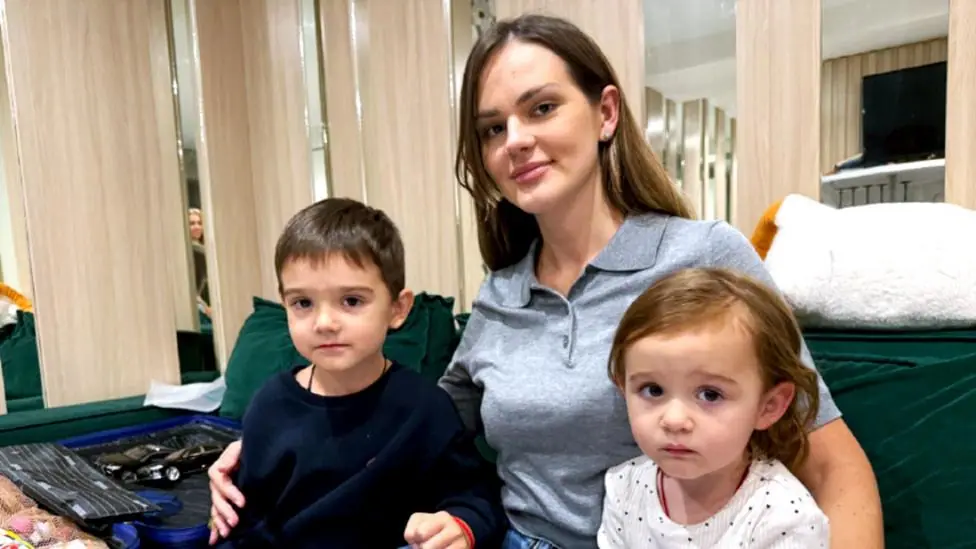 A woman with long brown hair sits on a green sofa with two young children, a boy and a girl.