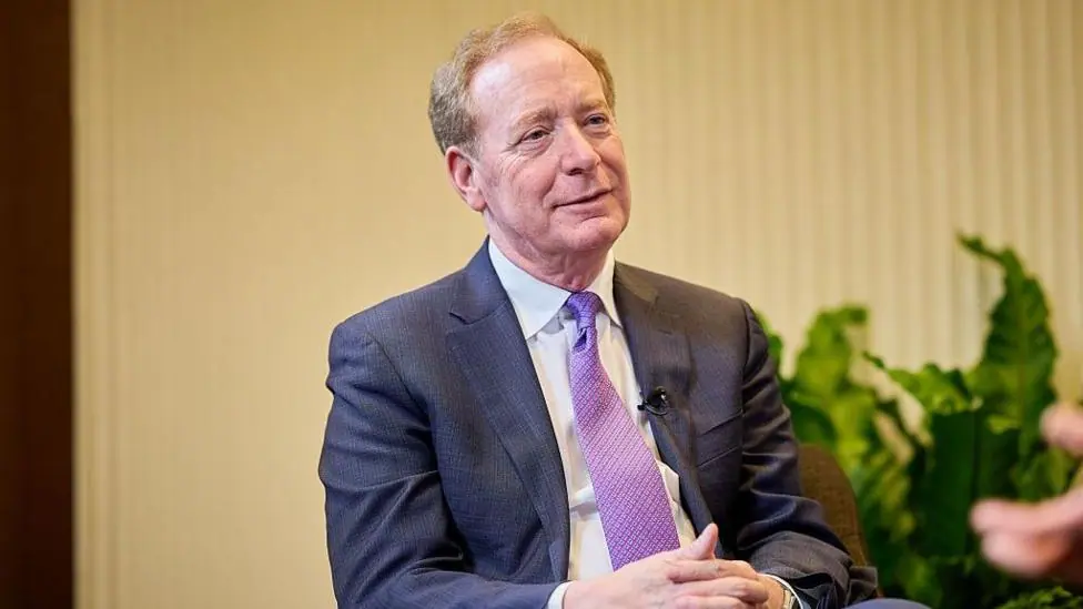 Microsoft president Brad Smith sitting with his hands clasped in his lap wearing a blue suit, white shirt, and purple-patterned tie. Behind him in soft focus, there is a beige background and some…