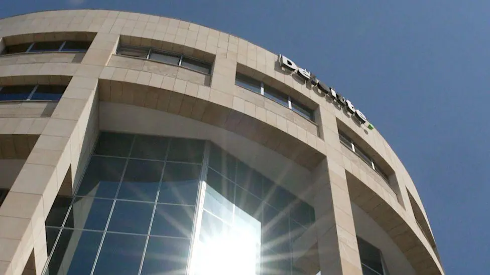 A view of Deloitte's offices in Limassol, Cyprus, looking up at a tall, curved building in honey-coloured stone and tinted glass, with blue sky above and a sunburst of reflected light at the bottom…