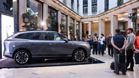 Side view of a gray Xpeng G9L SUV on a display platform in a courtyard with visitors gathered nearby.