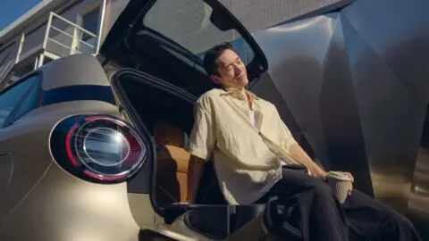 A man sitting on the lowered rear tailgate of a Smart #2 with the upper glass hatch open.