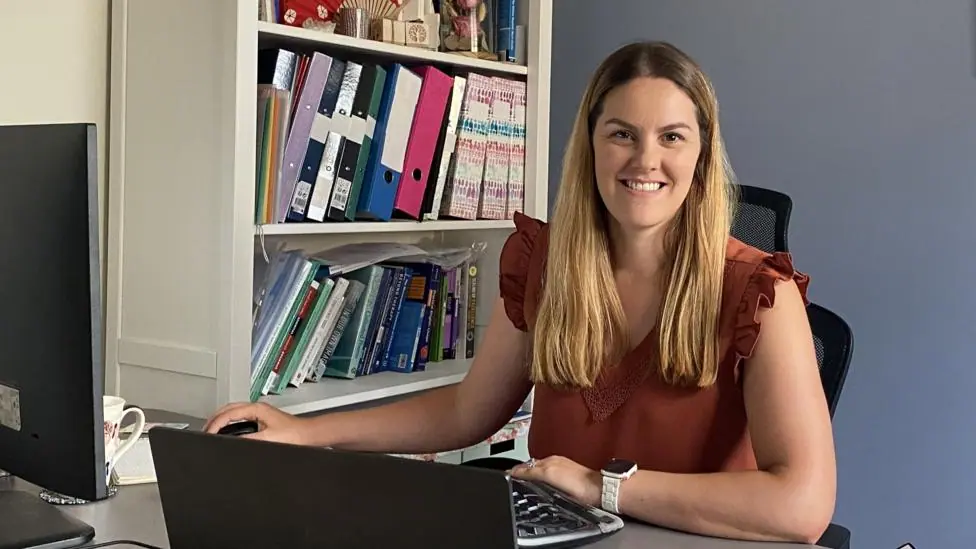 With long blonde hair and wearing a red short sleeve blouse, Carol Bereuter sits at her desk with a bookshelf to her right.