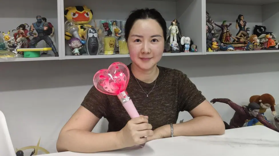 Melanie Wang from the Mihox sales team holds up a toy. She is sitting in front of shelves of other toys