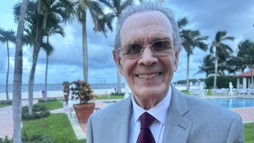 Juan Omar Sixto stands in front of a swimming pool and Biscayne Bay in Florida. He is smartly dressed in a suit and tie
