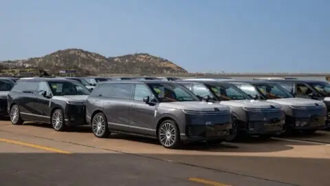 A silver Xpeng GX SUV parked in front of rows of vehicles with protective coverings at a waterfront terminal.