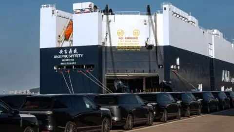 Rows of Xpeng GX SUVs parked in front of a vehicle carrier ship with its loading ramp lowered.