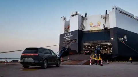 A Xpeng GX SUV approaches a vehicle carrier ship's loading ramp as port workers stand nearby.