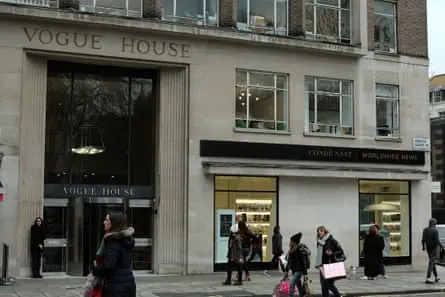 People walk past the frontage of Vogue House, a stone building with an imposing glass doorway and revolving doors. The scene appears wintry with the passersby wearing hats, coats and scarves, and…