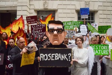 A protester in a Burnham mask holds a “Rosebank?” sign amid activists dressed as devils and angels