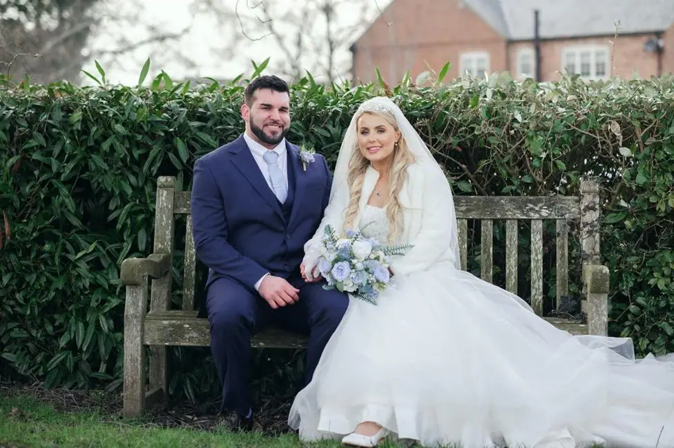 Simon Greenwood and Rosey Greenwood sitting on a bench on their wedding day, with Simon in a suit and Rosey in a wedding dress