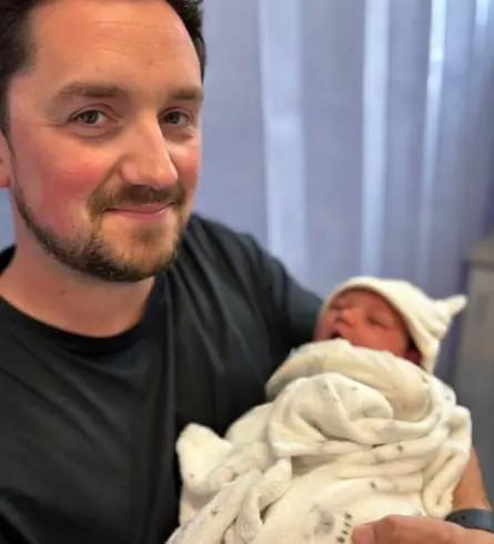 Joe Erskine smiles while holding his newborn baby wrapped in a blanket