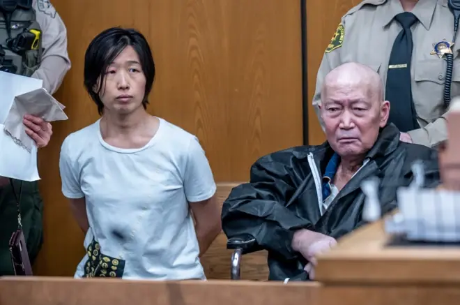 張莎莎身穿白色T恤、雙手放在身後，在法庭內坐在宣國軍旁邊。宣國軍是一名禿頭的中年男子，兩人身後各有執法人員站立。