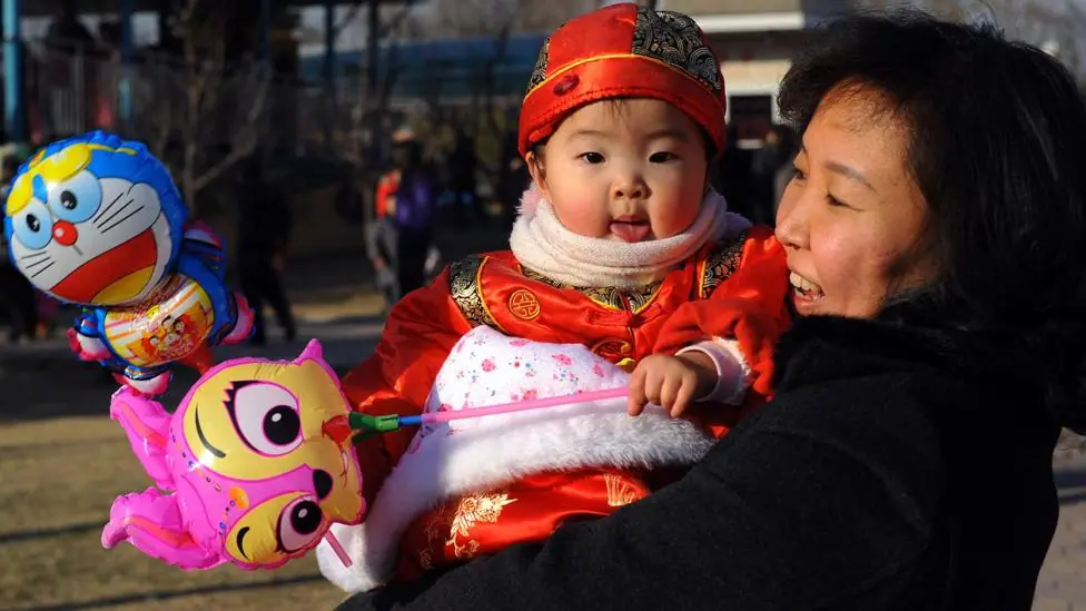 A Chinese woman is outside. She is laughing and holds a baby girl wearing traditional Chinese dress who she is looking at. The baby is holding a balloon