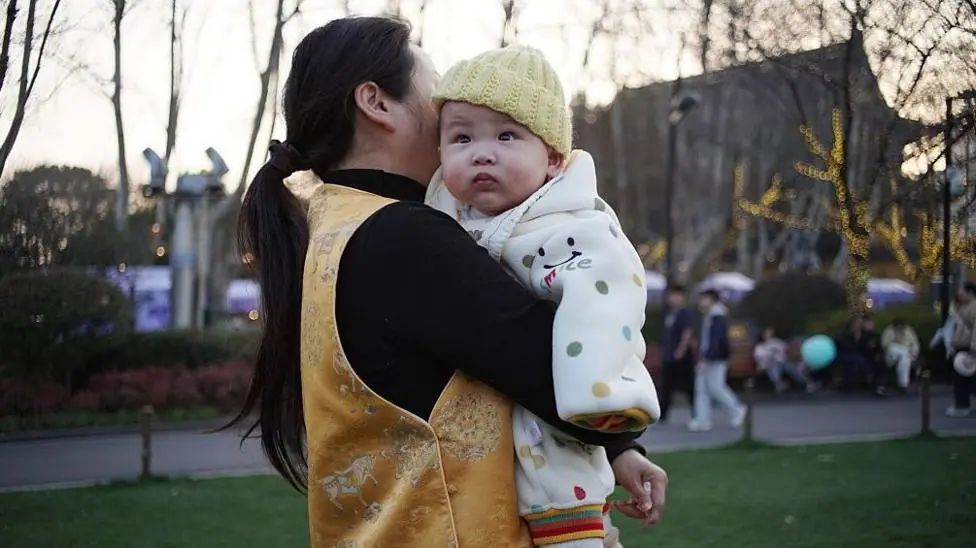 A woman in a golden vest holds a baby wrapped in a polka-dot blanket in the park at dusk